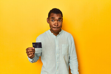 African American man with credit card, yellow studio, confused, feels doubtful and unsure.