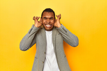 African American man in casual blazer, yellow studio, covering ears with hands.