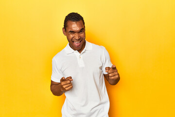 African American man in white polo, yellow studio, pointing to front with fingers.