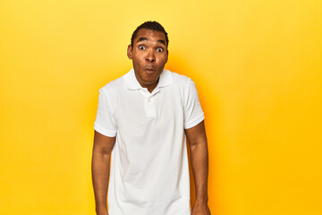 African American man in white polo, yellow studio, shrugs shoulders and open eyes confused.