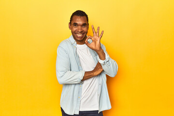 African American man in blue shirt, yellow studio, winks an eye and holds an okay gesture with hand.
