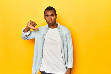 African American man in blue shirt, yellow studio, showing a dislike gesture, thumbs down....
