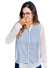 Beautiful hispanic woman wearing casual striped shirt smelling something stinky and disgusting,...
