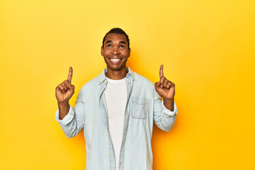 African American man in blue shirt, yellow studio, indicates with both fore fingers up showing a...