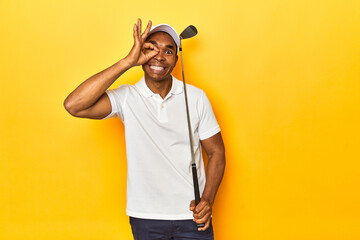 African American man golfer, yellow studio backdrop, excited keeping ok gesture on eye.