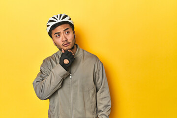 Asian man in bike helmet and gloves, yellow studio backdrop looking sideways with doubtful and skeptical expression.