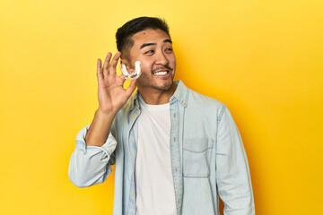 Asian man with invisible dental corrector, yellow studio backdrop looks aside smiling, cheerful and...