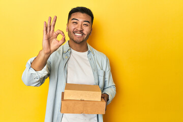 Asian man holding delivery food boxes, yellow studio backdrop cheerful and confident showing ok...