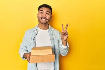 Asian man holding delivery food boxes, yellow studio backdrop showing number two with fingers.
