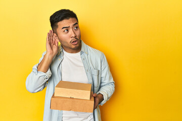 Asian man holding delivery food boxes, yellow studio backdrop trying to listening a gossip.