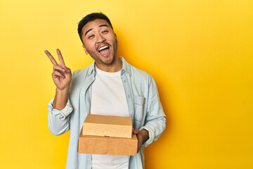 Asian man holding delivery food boxes, yellow studio backdrop joyful and carefree showing a peace...