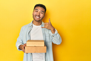 Asian man holding delivery food boxes, yellow studio backdrop showing a mobile phone call gesture...