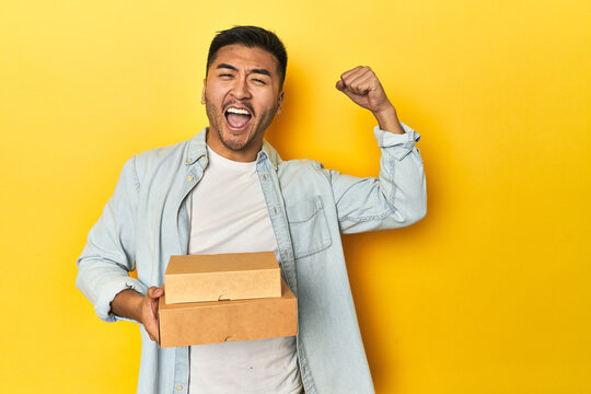 Asian man holding delivery food boxes, yellow studio backdrop raising fist after a victory, winner concept.