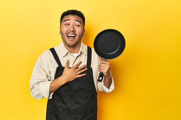 Asian chef holding a pan, yellow studio backdrop laughs out loudly keeping hand on chest.