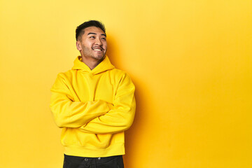 Bold Asian man in yellow hoodie on yellow studio smiling confident with crossed arms.