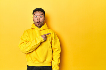 Bold Asian man in yellow hoodie on yellow studio pointing to the side