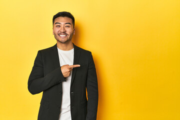 Asian man in black business jacket on yellow studio smiling and pointing aside, showing something...