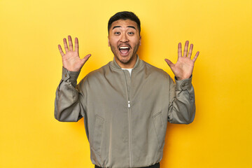 Stylish Asian man in gray jacket on yellow studio receiving a pleasant surprise, excited and...