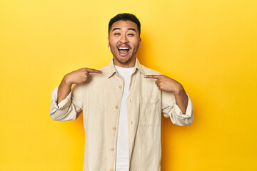 Casual Asian man with open shirt, white tee on yellow studio surprised pointing with finger,...