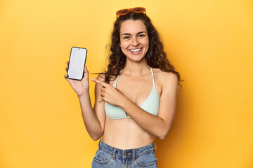 Young Caucasian woman in bikini showing mobile screen in studio with yellow backdrop. - obrazy, fototapety, plakaty
