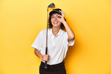 Athletic Caucasian woman with curly hair golfing in studio excited keeping ok gesture on eye.