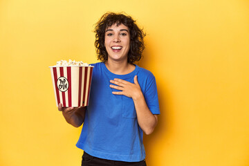 Curly-haired woman with popcorn for movie, studio laughs out loudly keeping hand on chest.