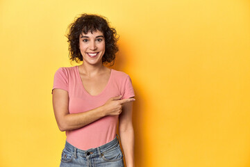 Curly-haired Caucasian woman in pink t-shirt smiling and pointing aside, showing something at blank space.