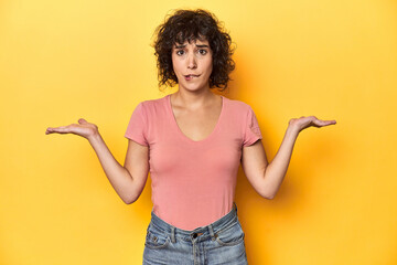 Curly-haired Caucasian woman in pink t-shirt confused and doubtful shrugging shoulders to hold a...