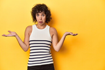 Caucasian curly-haired woman in white tank-top doubting and shrugging shoulders in questioning...