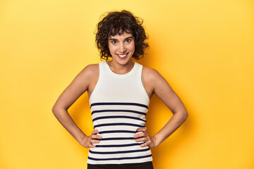 Caucasian curly-haired woman in white tank-top confident keeping hands on hips.