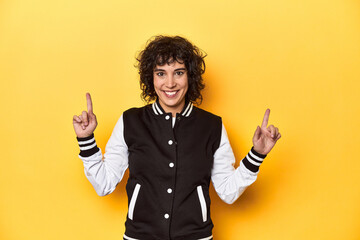 Curly-haired Caucasian woman in baseball jacket indicates with both fore fingers up showing a blank...