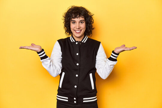 Curly-haired Caucasian woman in baseball jacket makes scale with arms, feels happy and confident.