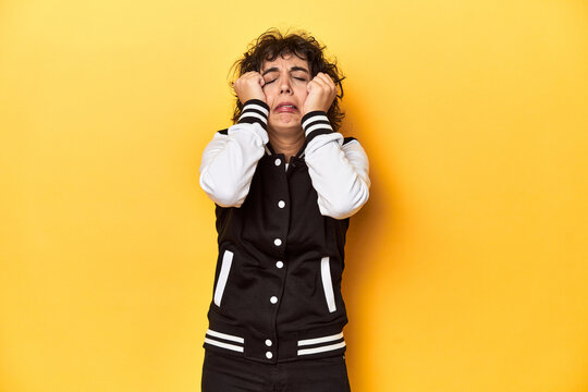 Curly-haired Caucasian woman in baseball jacket crying, unhappy with something, agony and confusion concept.