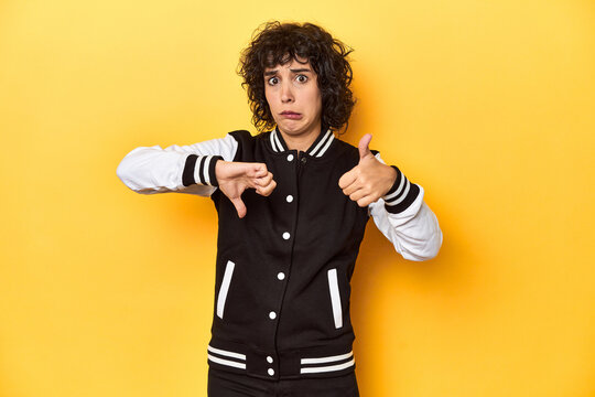 Curly-haired Caucasian woman in baseball jacket showing thumbs up and thumbs down, difficult choose concept