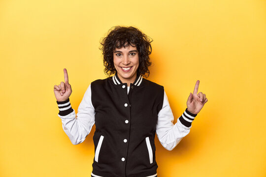 Curly-haired Caucasian woman in baseball jacket indicates with both fore fingers up showing a blank space.