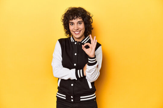 Curly-haired Caucasian woman in baseball jacket winks an eye and holds an okay gesture with hand.