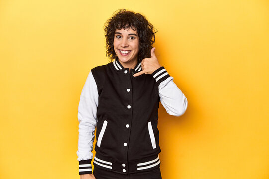 Curly-haired Caucasian woman in baseball jacket showing a mobile phone call gesture with fingers.