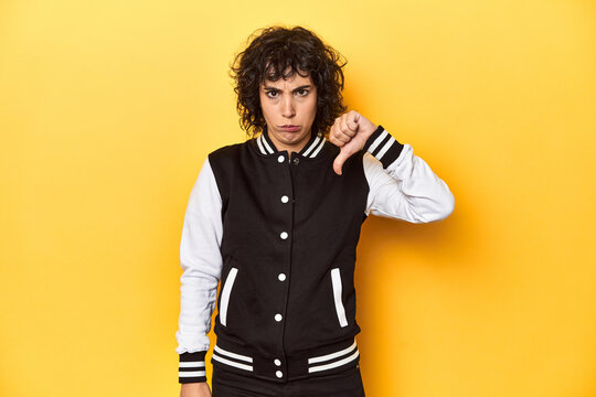 Curly-haired Caucasian woman in baseball jacket showing a dislike gesture, thumbs down. Disagreement concept.