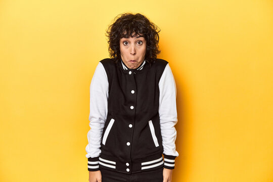 Curly-haired Caucasian woman in baseball jacket shrugs shoulders and open eyes confused.