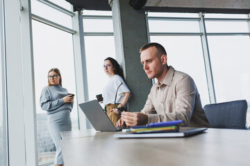 A young man works in the office on a laptop while sitting at the table against the background of colleagues. Modern workspace with office workers.