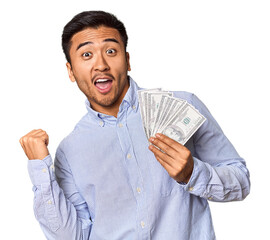 Young Chinese man holding dollar bills in studio points with thumb finger away, laughing and...