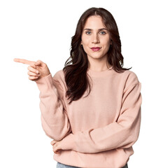 Young Caucasian woman in a studio setting smiling cheerfully pointing with forefinger away.