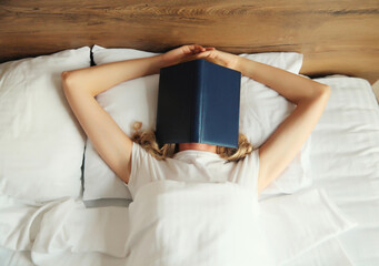 Young sad tired woman student covering her face with open book with blank cover while lying on the...