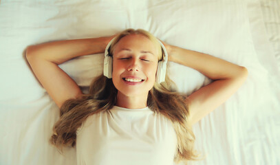 Happy relaxed middle-aged woman listening to music with wireless headphones while lying on bed in bedroom at home