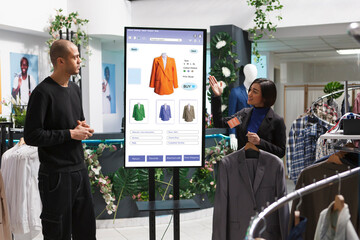 Asian store assistant showcasing items to man in front of self service kiosk, selecting fashion collection clothes from clothing boutique. Caucasian customer discussing products with female employee.