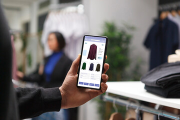 Caucasian customer in a clothes store is holding a smartphone and looking at clothes on a boutique website. Male shopaholic is perusing clothing items in a mall using a cellphone.