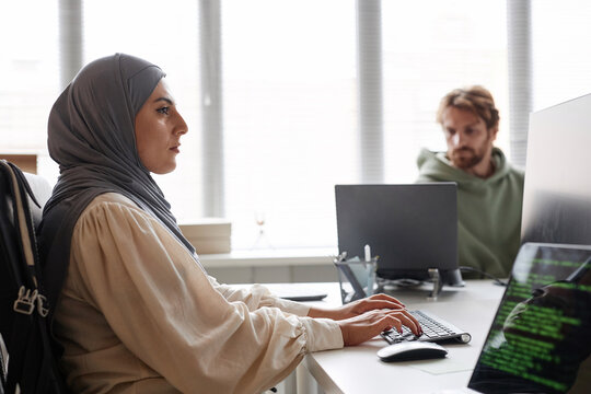Side view portrait of young Middle-Eastern woman as female programmer using computer in office and wearing headscarf