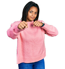 Beautiful hispanic woman wearing casual winter sweater punching fist to fight, aggressive and angry attack, threat and violence