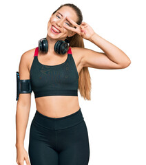 Beautiful blonde woman wearing gym clothes and using headphones doing peace symbol with fingers over face, smiling cheerful showing victory