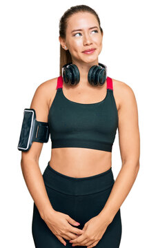 Beautiful blonde woman wearing gym clothes and using headphones smiling looking to the side and staring away thinking.